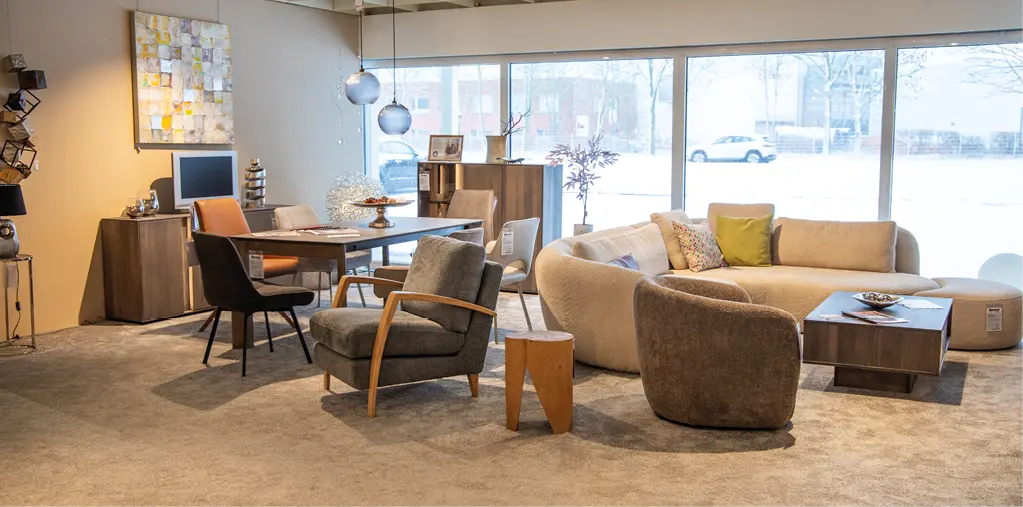 Ausstellung mit hellem Sofa, Sesseln und kleinem Tisch rechts, links Essbereich mit Holztafel und Stühlen, große Fensterfront im Hintergrund und Wanddeko.